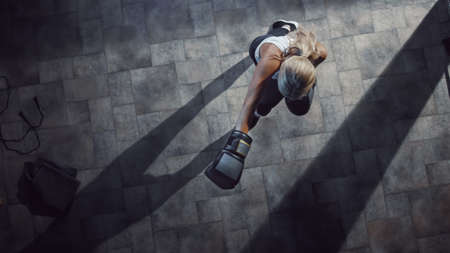 Professional Female Boxer Exercises Air Punches while Wearing Sportswear and Fighting Gloves in the Hardcore Gym. Strong Beautiful Girl Kickboxing and Self Defense Workout. Top Dramatic Viewの写真素材