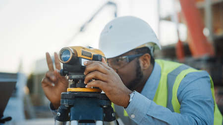On the Commercial Industrial Building Construction Site: Professional Engineer Surveyor Takes Measures with Theodolite, Worker Uses Laptop. In the Background Skyscraper Formwork Frames and Craneの写真素材
