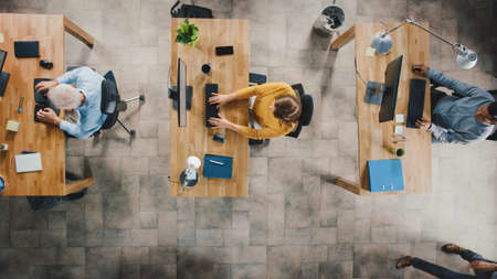 Top View Shot in the Office: Diverse Group of Business Startup Employees Sitting at their Desks Working on Desktop Computers. Stylish and Modern Office Loft with Professionals Doing their Jobの写真素材