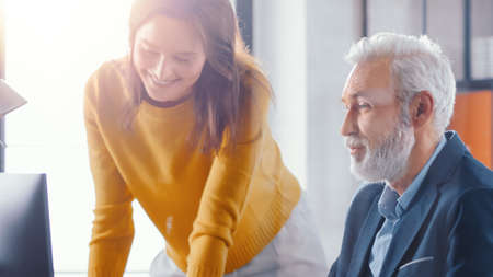 Handsome Middle Aged Creative Engineer Sitting at the Desk working on Desktop Computer Has Discussion with Beautiful Female Project Manager. Office with Friendly Colleagues Chatting, Finding Solutionsの写真素材