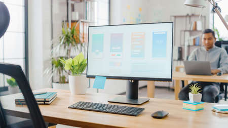 Desktop Computer Standing on the Desk in the Modern Creative Office. Screen Showing Smartphone Application UI UX. In the Background Bright Studio with Young Professionals Work on Computers.の写真素材