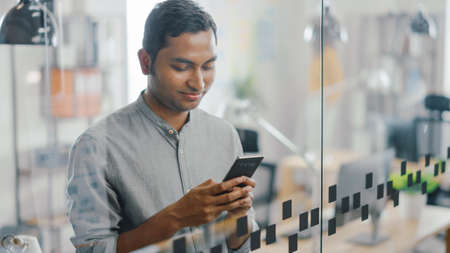 Portrait of Handsome Professional Indian Man Uses Mobile Phone, Writes Important Email. Successful Man Using Smartphone Working in Bright Diverse Office.の写真素材