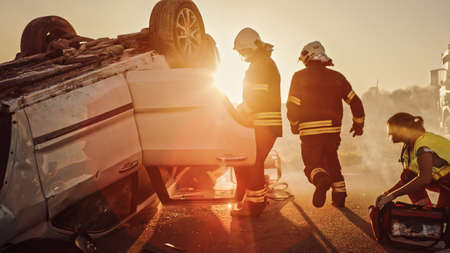 Car Crash Traffic Accident: Paramedics and Firefighters Plan Rescuing Passengers Trapped in Rollover Vehicle. Medics Prepare Stretchers and First Aid Equipment. Firemen Use Hydraulic Cutters Spreaderの写真素材