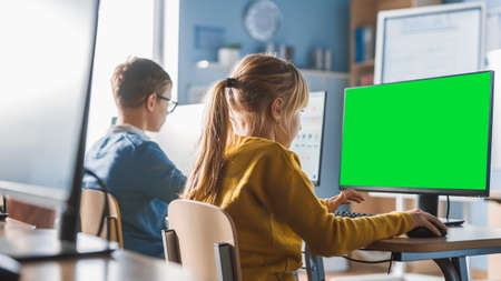 Elementary School Computer Science Classroom: Cute Little Girl Uses Green Mock-up Screen Computer while Learning Coding and Programming. Schoolchildren Getting Modern Education. Over the Shoulderの写真素材
