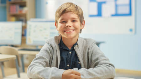 Portrait of a Cute Little Boy Sitting at his School Desk, Smiles Happily. Smart Little Boy with Charming Smile Sitting in the Classroom.の写真素材