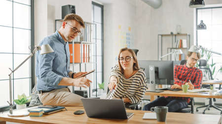 Male Colleague Dominantly Sits on a Table in Front of a Young Female Cowroker while Holding a Tablet...