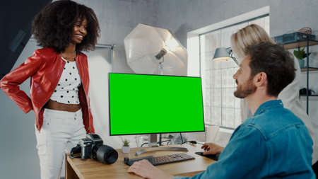 In Photo Studio Fashion Magazine Designer and Beautiful Black Cover Girl talk to a Professional Photographer who Uses Desktop Computer with Green Mock-up Screen Displayの写真素材
