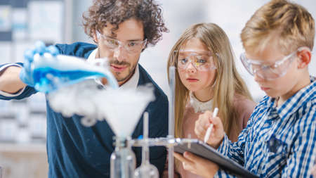 Elementary School Science Classroom: Enthusiastic Teacher Explains Chemistry to Diverse Group of Children, Shows them How to Mix Chemicals in Beakers. Children Use Digital Tablet Computers and Talkの写真素材