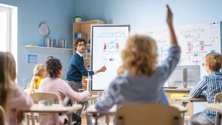 Brilliant Schoolboy Listens Attentively to His Teacher Explaning Lesson and Raises His Hand with a Question. In Elementary School with Group of Bright Multiethnic Kids Learning Science. Back View Shotの写真素材