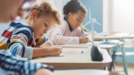 In Elementary School Classroom Brilliant Caucasian Boy Writes in Exercise Notebook, Taking Test and Writing Exam. Junior Classroom with Group of Children Working Diligently and Learning New Stuffの写真素材