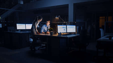 Ambitious and Confident Businessman Uses Desktop Computer, His Female Project Manager Explains Specific Tasks, Account Handling and Strategic Moves. Professional People Late at Night in Big Officeの写真素材