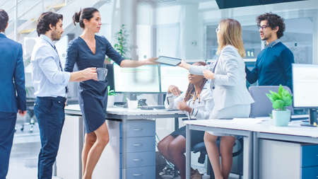 In the Bright Busy Office Group of Young Professionals have Team Building Meeting. Standing Around the Desk They Discuss Problem Solutions, Chat, Plan and Strategics, Pass Digital Tabletsの写真素材