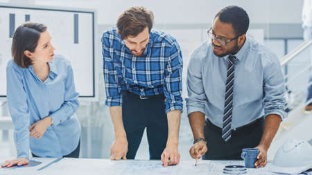 In the Industrial Engineering Facility: Diverse Group of Engineers and Technicians on a Meeting Gather Around Table Unravel Sheets of Engine Design Technical Drafts, Have Discussion, Analyse Drawingsの写真素材