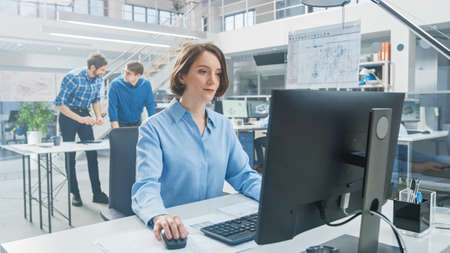 In the Industrial Engineering Facility: Portrait of the Smart and Beautiful Female Engineer Working on Desktop Computer. In the Background Specialists and Technicians Working with Drafts and Drawingsの写真素材