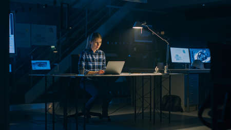 Beautiful Female Engineer Sitting at Her Desk Works on a Laptop Computer. Blueprints Lying on a...
