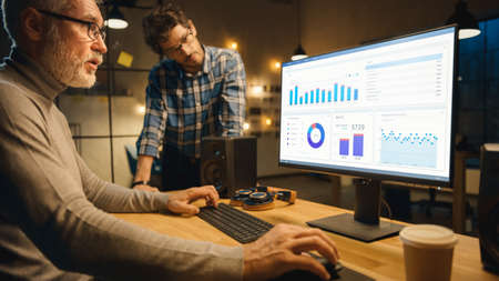 Professional Senior Business Martket Analyst Works on Desktop Computer Working with Charts, Graphs, Has Conversation with Younger Specialist who Gives Him Advice. Modern Business Office in Eveningの写真素材