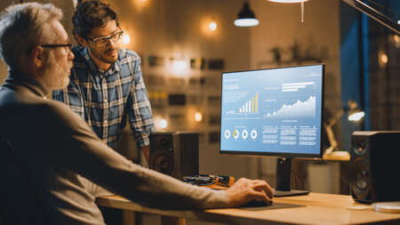 Professional Senior Business Martket Analyst Works on Desktop Computer Working with Charts, Graphs, Has Conversation with Younger Specialist who Gives Him Advice. Modern Business Office in Eveningの写真素材