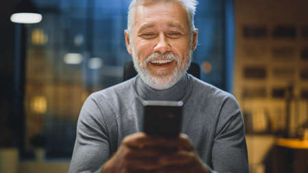 Portrait of Handsome and Successful Middle Aged Bearded Businessman Uses Smartphone Laughs and Smiles. Working from Cozy Home Office Studioの写真素材