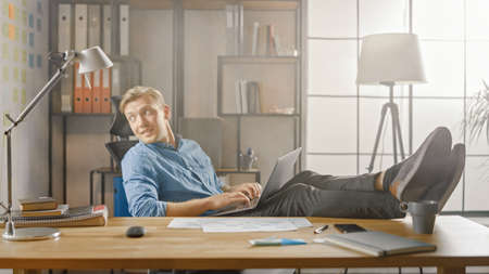 Handsome Young Entrepreneur Works on a Laptop with His Legs Up on the Desk. Designer Uses Notebook...