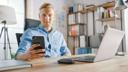 Businessman Sitting at His Desk Works on Desktop Computer in the Stylish Office, Picks up and Starts Using Smartphone, Uses Social Media App, Emailing Business Partners, Messagingの写真素材