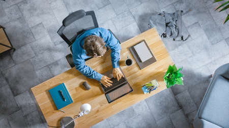 Top View Shot: Businessman Sitting at the Wooden Desk Works on a Laptop in his Home Office. He Types, Writes, Surfs the Internet, Creates and Designs Software, Online Shoppingの写真素材