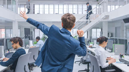 Back View Shot of a Young Cheerful Handsome Business Manager Wearing a Suit and Tie Dancing in the Office. Diverse and Motivated Business People Work on Computers in Modern Open Office.の写真素材