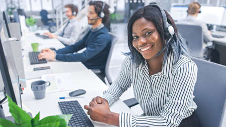 Beautiful Young Female Customer Service Operator Smiling for a Portrait at a Busy Modern Call Center with Diverse Multicultural Team of Office Specialists Wearing Headsets and Taking Calls.の写真素材