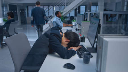 Young Japanese Manager is Tired at his Work Place with Desktop Computer and Falls Asleep. Diverse...