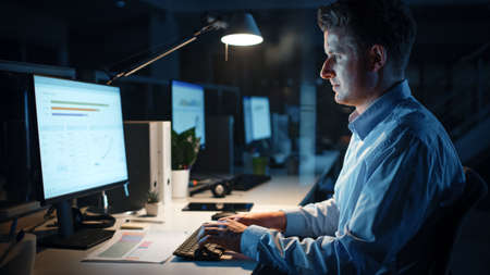 Portrait of Ambitious and Confident Businessman Uses Desktop Computer, Typing on a Keyboard, Monitoring Business Transactions, Signing Contracts. Works Late at Night in Big Corporate Officeの写真素材