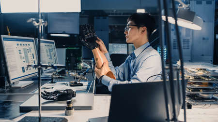Futuristic Prosthetic Robot Arm Being Tested by a Professional Japanese Development Engineer in a High Tech Research Laboratory with Modern Computer Equipment. He is Satidfied with the Result.の写真素材