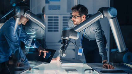 Diverse Team of Engineers with Laptop and a Tablet Analyse and Discuss How a Futuristic Robotic Arm Works and Moves a Metal Object. They are in a High Tech Research Laboratory with Modern Equipment.の写真素材