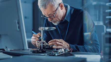 Electronics Maintenance Engineer Soldering Motherboard, Microchip and Circuit Board, Looking through Magnifying Glass, Consults Personal Computer. Electronics Repair and Testingの写真素材