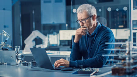 Professional Electronics Design Engineer Wearing Glasses Works on Computer in Research Facility. In the Background Motherboards, Circuit Boards and Computer Components in the High Tech Facility.の写真素材