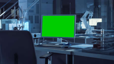 On the Desk Computer with Isolated Green Mock-up Screen Display. In the Background Robot Arm Concept Standing in Heavy the Dark.Industry Engineering Facilityの写真素材