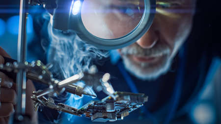 Electronics Maintenance and Repair Engineer Soldering Motherboard, Microchip and Circuit Board, Looking through Magnifying Glass, Consults Personal Computer. Close-up Low Angle Shotの写真素材