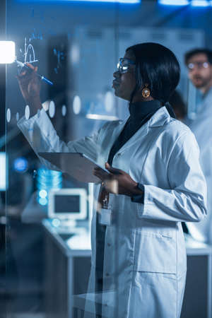 In the Research Laboratory Smart and Beautiful African American Female Scientist Wearing White Coat and Protective Glasses Writes Formula on Glass Whiteboard, References Her Tablet Computerの写真素材