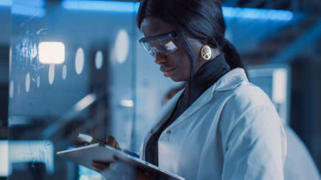 In the Research Laboratory Smart and Beautiful African American Female Scientist Wearing White Coat and Protective Glasses Writes Formula on Glass Whiteboard, References Her Tablet Computerの写真素材