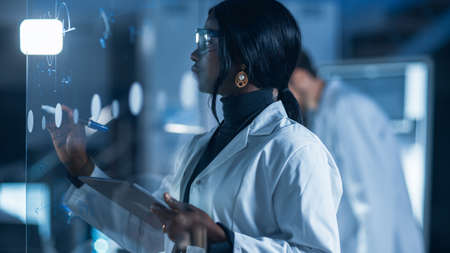 In the Research Laboratory Smart and Beautiful African American Female Scientist Wearing White Coat and Protective Glasses Writes Formula on Glass Whiteboard, References Her Tablet Computerの写真素材