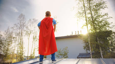 Boy is Playing a Role of a Super Hero. Hes Standing on a Roof of a House with His Hands on His...