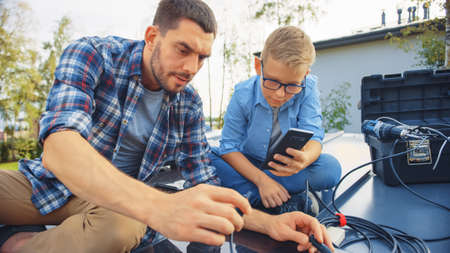 Father and Son Installing Solar Panels to a Metal Basis. They Work with Wiring on a House Roof on a Sunny Day. Boy is Looking at a Manual on a Smartphone. Father Connects the Plugs.の写真素材