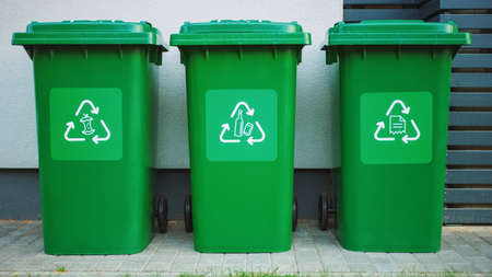 Green colored, plastic garbage bins, with different recycle logos on front, stacked in row against...