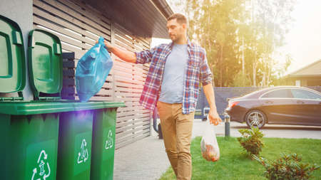 Caucasian Man is Throwing Away Two Plastic Bags of Trash next to His House. One Garbage Bag is Sorted with Biological Food Waste, Other with Recyclable Bottles Garbage Bin.の写真素材