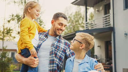Portrait of a Happy Family of Three: Father, Daughter, Son. They Are Posing In Front of Camera on a Lawn Next to Their Country House. Dad is Holding the Girl in His Arms. Boy is Holding a Football.の写真素材