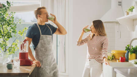 Handsome Young Man in Glasses Wearing Apron and Beautiful Girl are Making A Smoothie in the Kitchen. Happy Couple are Trying Healthy Organic Beverage. Male and Female at Home on a Sunny Day.の写真素材