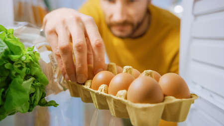Shot from Inside Kitchen Fridge: Handsome Man Opens Fridge Door, Looks inside Takes Few Eggs from Eggs Box. Man Preparing Healthy Meal. Point of View POV Shot from Refrigerator full of Healthy Foodの写真素材
