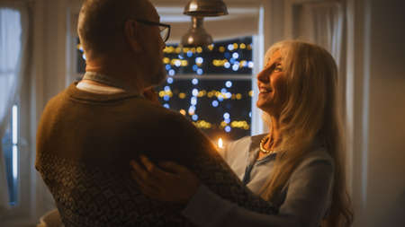 Senior Couple in Love Have Romantic Evening, Dancing in the Dining Room, Celebrating Anniversary. Portrait of a Happy Elderly Husband and Wife Have Lovely Evening with Festive Table in Cozy Kitchenの写真素材