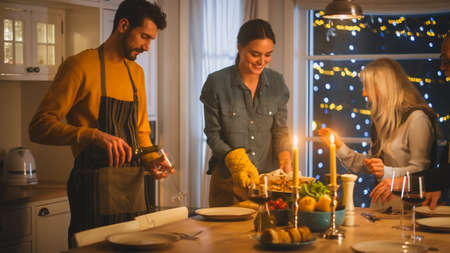 Multi Generation Family of Celebrating together, Gathering around the Table with Delicious Dinner Meal. Young Mother Takes Dish out of Oven Grandparents Admire How Delicious Casserole Looksの写真素材