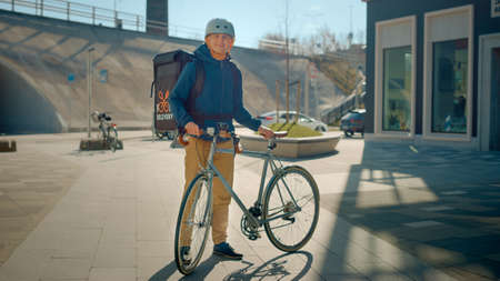 Handsome Happy Food Delivery Man Wearing Thermal Backpack and Safety Helmet Stands Beside his Bike...