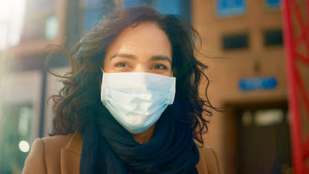 Portrait of a Beautiful Dark Haired Young Woman Wearing Protective Medical Face Mask and Standing on the Street. Safe and Happy Woman Practicing Social Distancing, Quarantine. Blurred City Backgroundの写真素材