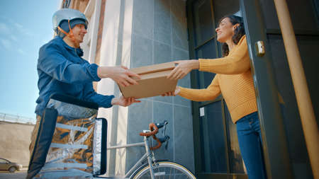 Happy Food Delivery Man Wearing Thermal Backpack on a Bike Delivers Pizza Order to a Beautiful Female Customer. Courier Delivers Takeaway Fast Food to a Smiling Girl in Office Building. Low Angleの写真素材